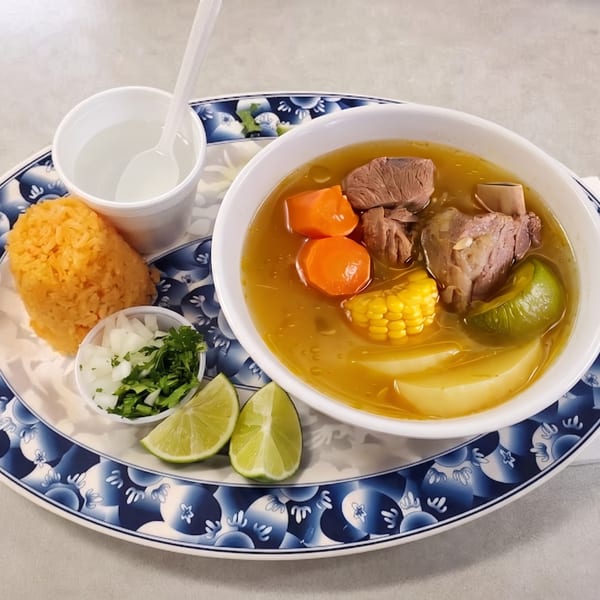 Hearty beef soup with corn, carrots, and potatoes, served with rice, onions, cilantro, and lime.