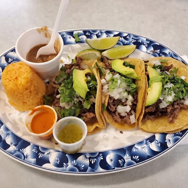 Three street tacos with meat, onions, cilantro, and avocado, served with rice, lime, and sauces.
