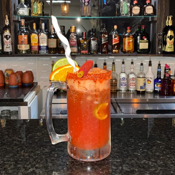 A large red cocktail with a chili straw and citrus slices sits on a dark bar counter.